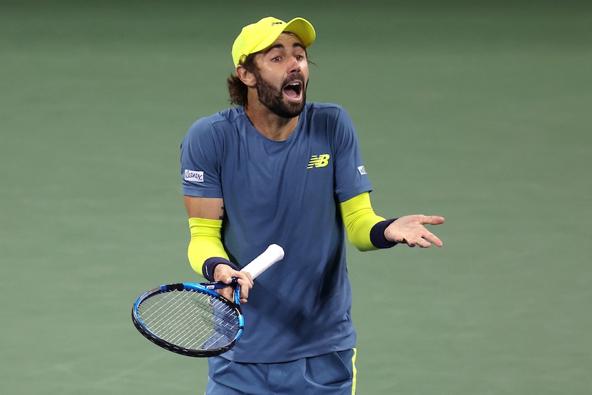 A tennis player wearing a yellow cap, T-shirt and shorts holds a racket in one right hand and gesticulates with the other.